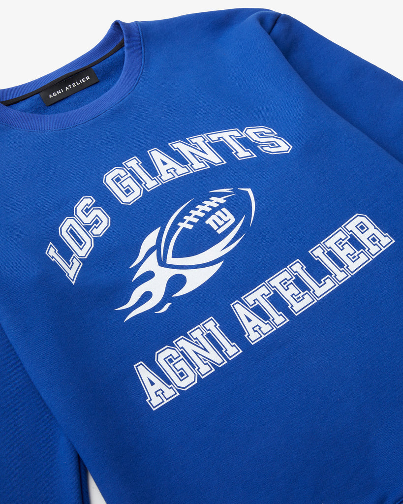 An angled artistic shot of a Blue Crew Neck with Los GIANTS on it, featuring a graphic of a football on fire with NY GIANTS and the Agni Atelier logo.