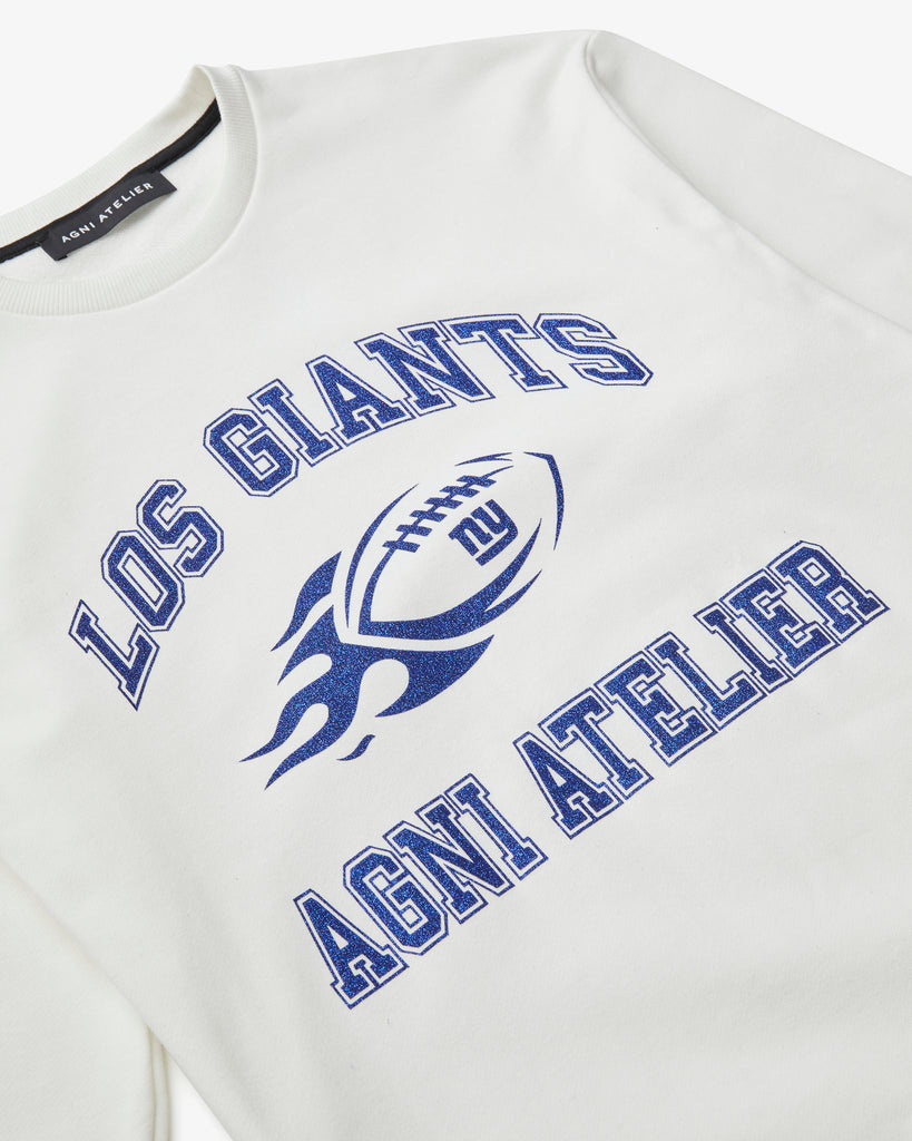 An artistic shot of a White Crew Neck with Los GIANTS on it, featuring a graphic of a football on fire with NY GIANTS and the Agni Atelier logo. The Text and graphics all have a shimmering blue color.