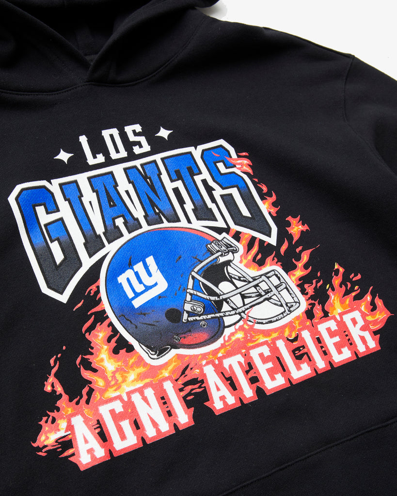 An angled artistic shot of a Black Hoodie with Los GIANTS emblazoned upon it, featuring a graphic of the NY GIANTS NFL FOOTBALL TEAM HELMET and the Agni Atelier logo in the foreground, with flames in the background