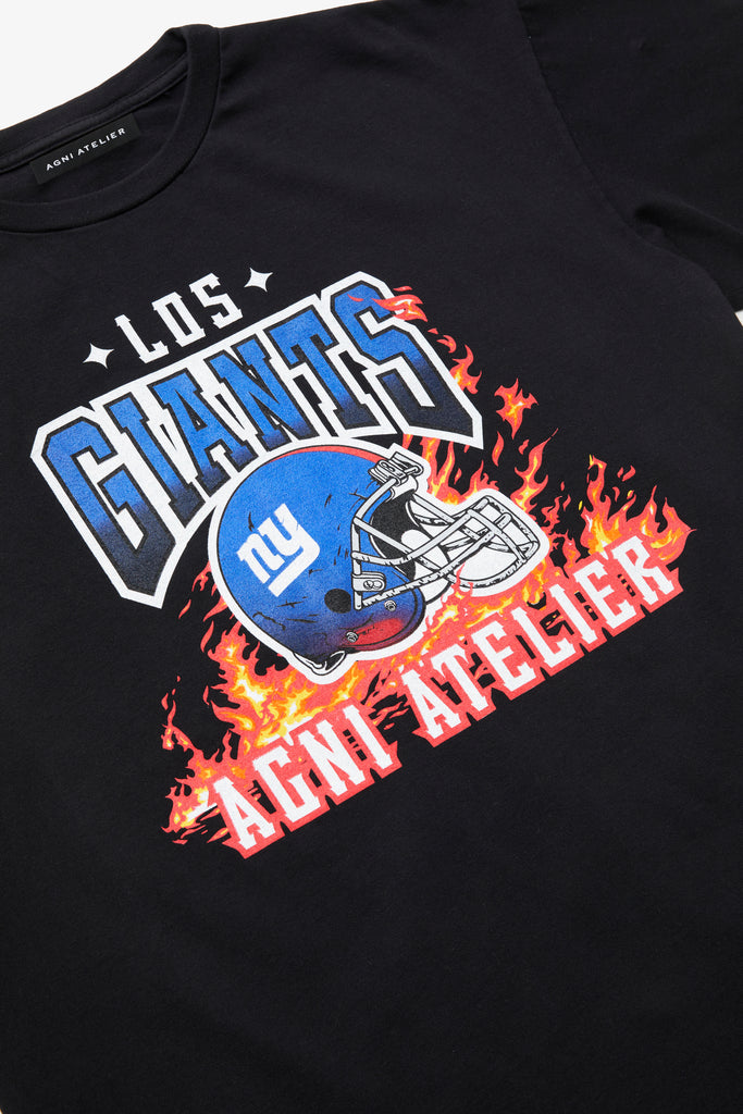 An angled artistic shot of a Black Tee Shirt with Los GIANTS emblazoned upon it, featuring a graphic of the NY GIANTS NFL FOOTBALL TEAM HELMET and the Agni Atelier logo in the foreground, with flames in the background